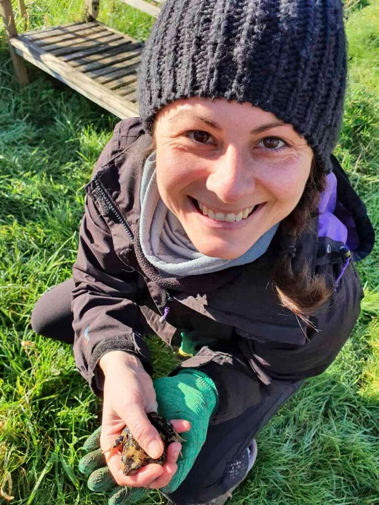 Donna sorridente con foulard scuro e felpa verde, in un ambiente naturale. Con una rana in mano