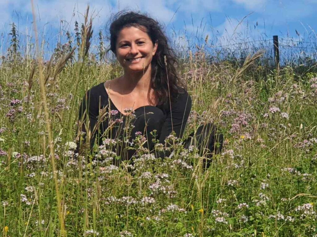Donna sorridente con capelli scuri e maglietta verde scuro, circondata da vegetazione.