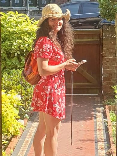 Donna in un abito rosso lungo, cammina o posa su una strada alberata con case sullo sfondo.