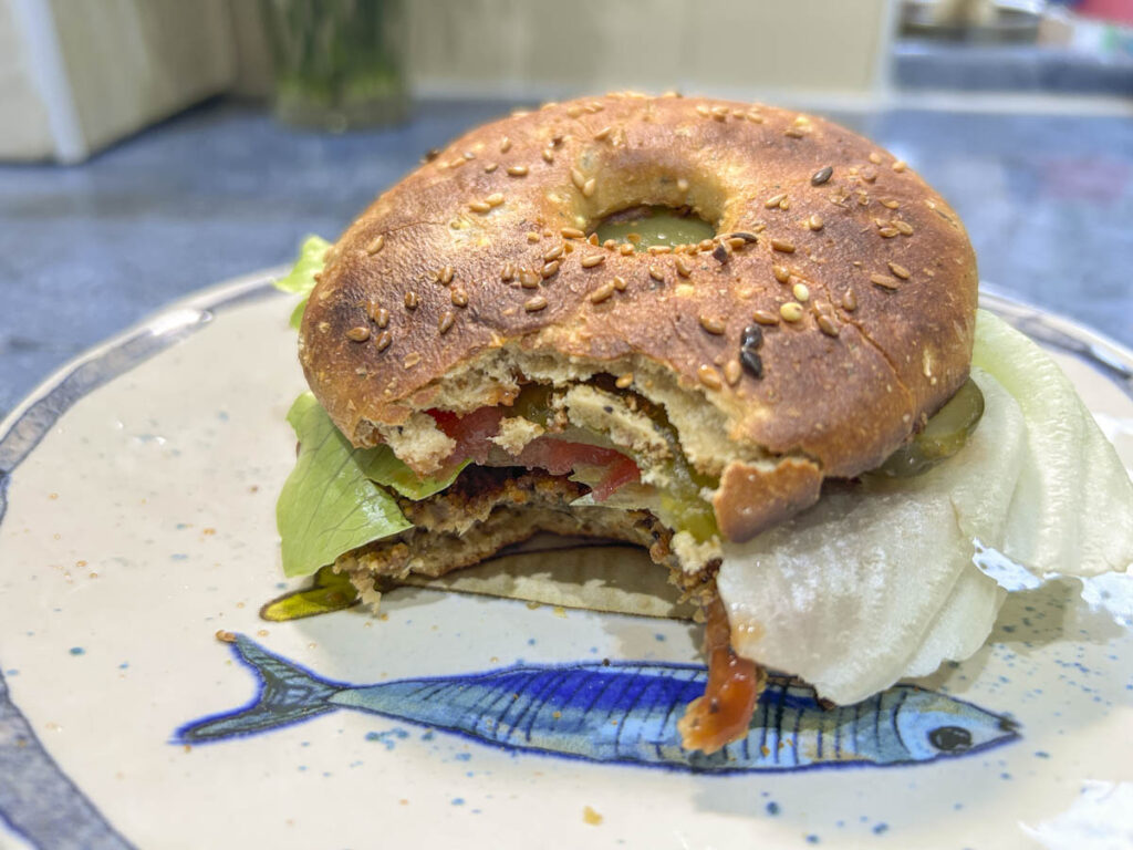 Hamburger vegano con funghi e avena servito in un bagel proteico con insalata, pomodoro e cetriolini.