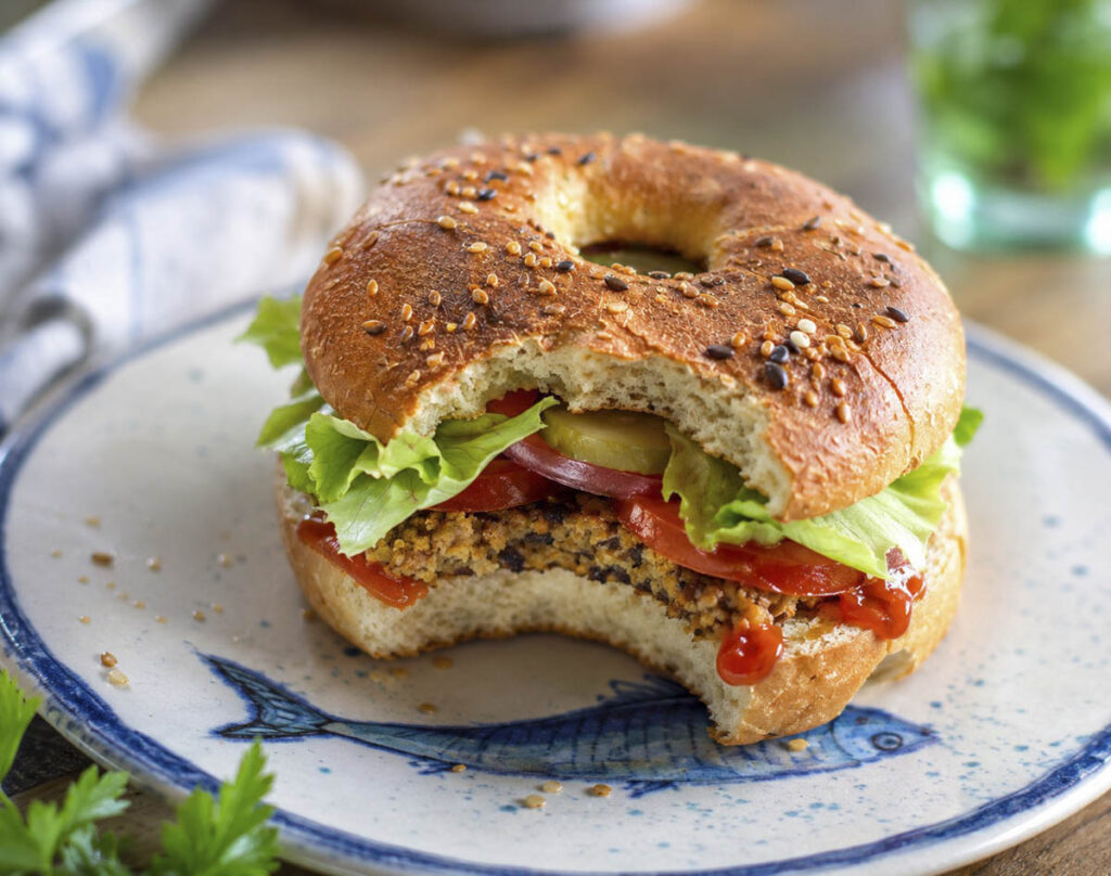 Hamburger vegano con funghi e avena servito in un bagel proteico con insalata, pomodoro e cetriolini.